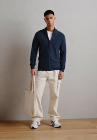 Suéter de punto azul marino con cremallera, pantalones cargo beige con bolsillos laterales, camiseta blanca y zapatillas. La modelo sostiene un bolso tote beige.
