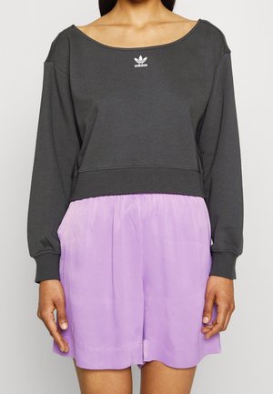 Kobieta w ciemnoszarej bluzie Adidas z dekoltem z odkrytymi ramionami oraz jasnofioletowych szortach z wysokim stanem.