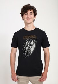 Sort bomulds T-shirt med et grafisk design af et ansigt med kløemærker. Teksten lyder "John Carpenters Halloween" med fed orange.
