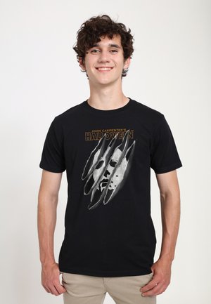 Sort bomulds T-shirt med et grafisk design af et ansigt med kløemærker. Teksten lyder "John Carpenters Halloween" med fed orange.