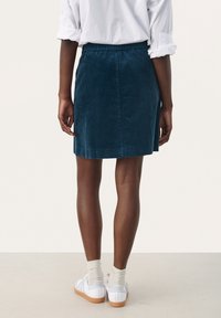 Falda de pana de color verde azulado oscuro con cintura elástica. De largo hasta la rodilla, textura suave, diseño sencillo, combinada con zapatillas blancas y calcetines.