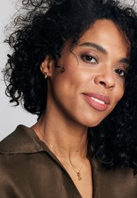 Złote kolczyki w kształcie serca oraz złoty naszyjnik z zawieszką w kształcie nieskończoności noszone z ciemno-brązową, teksturowaną koszulą.