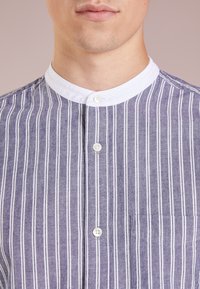 Camicia leggera con bottoni, a righe verticali blu e bianche, colletto bianco e una tasca sul lato sinistro.