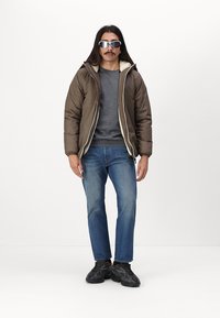 Giacca imbottita marrone con fodera in shearling, maglione grigio, jeans blu e sneakers nere, indossata da una persona in piedi contro uno sfondo bianco.