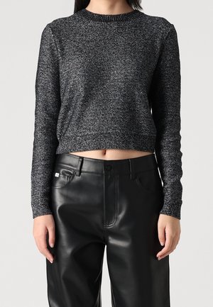 Vrouw draagt een langmouwige glinsterende donkergrijze cropped trui gecombineerd met een zwarte, hooggetailleerde leren broek, staand tegen een effen achtergrond.