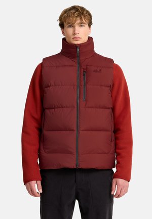 Mann trägt eine bordeauxrote Steppweste über einem roten, gerippten Langarmshirt und schwarzen Hosen, steht vor einem schlichten hellen Hintergrund.