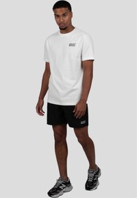 Camiseta blanca de algodón con un logo negro, combinada con pantalones cortos negros que tienen un pequeño logo. Llevada con zapatillas grises y negras.