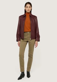 Donna in piedi che indossa una giacca di pelle bordeaux, un maglione a collo alto arancione, pantaloni kaki e stivali neri alla caviglia su uno sfondo neutro.