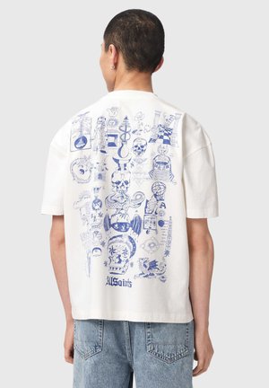 Person trägt weißes T-Shirt mit detailliertem blauem Tattoo-Motiv mit Schädeln, Schlangen, Sonne und mystischen Symbolen auf dem Rücken.