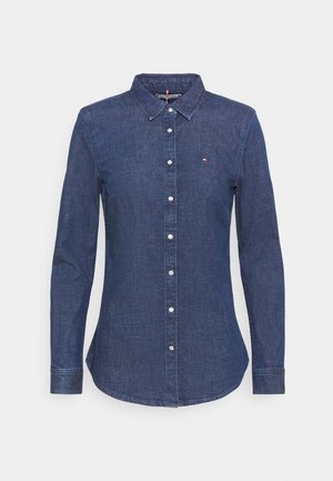 Chemise en denim bleu à manches longues, cintrée, avec col boutonné et boutons blancs, ornée d'un petit logo rouge, blanc et bleu sur la poitrine.