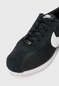 Crne tenisice s gornjim dijelom od velura i najlona, s bijelim Nike swoosh logom, crnim vezicama i strukturiranom bijelom gumenom petom.