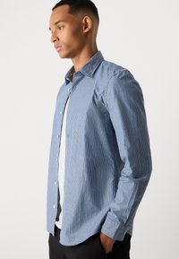 Cămașă cu dungi albastre, dintr-un material ușor, cu guler clasic și mâneci lungi, purtată peste un tricou gri deschis.