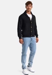 Cárdigan de punto oscuro con cuello de chal, botones de madera, llevado sobre una camiseta blanca y combinado con jeans de mezclilla azul claros y zapatillas blancas.