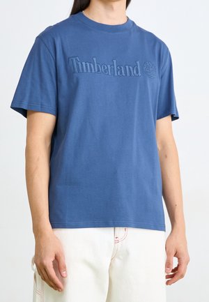 Sininen puuvillapaita, jossa on brodeerattu "Timberland" -logo ja puu-ikku, klassisella pyöreällä kaula-aukolla ja lyhyillä hihoilla.