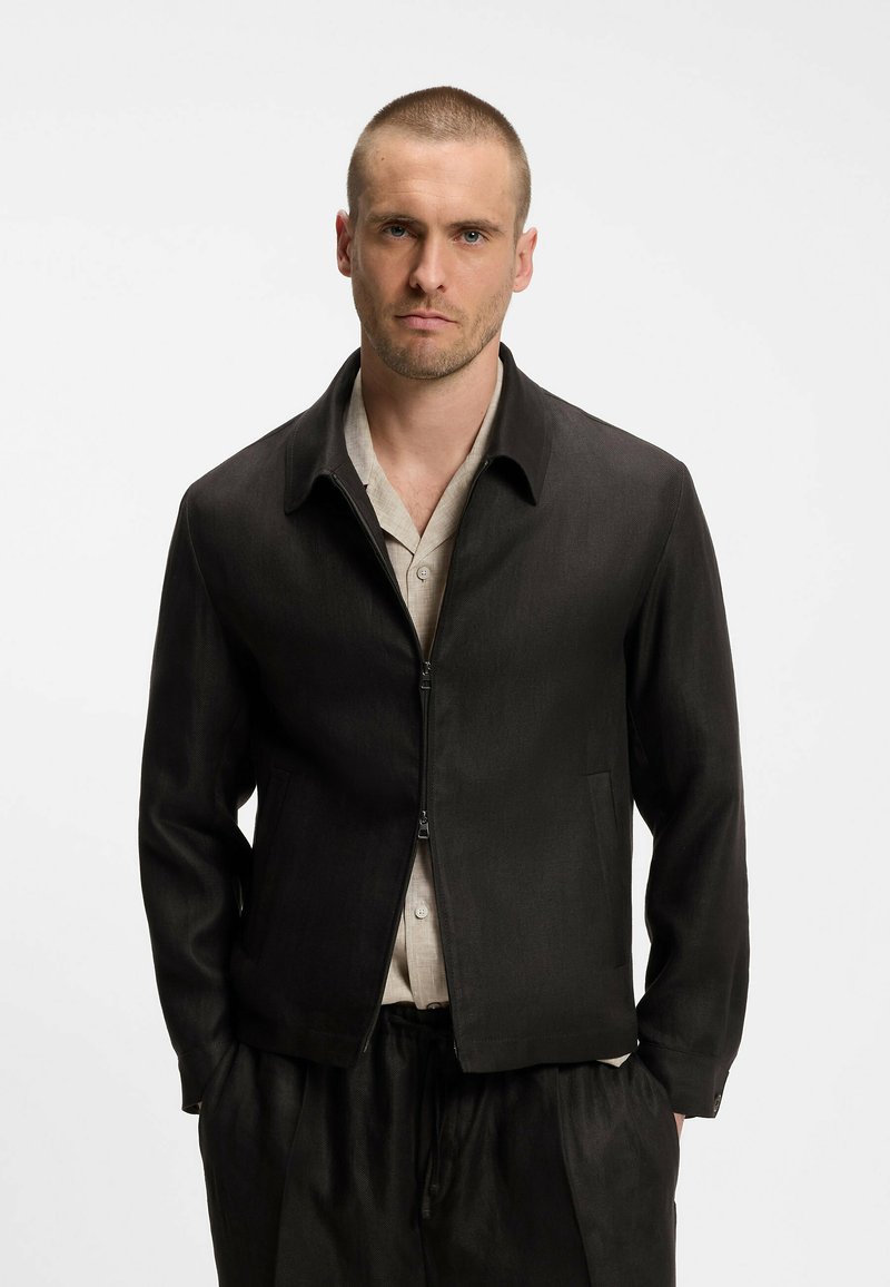 Homme aux cheveux courts portant une veste noire à fermeture éclair sur une chemise beige, debout les mains dans les poches devant un fond blanc uni.