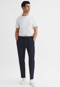 Camiseta blanca de cuello redondo, pantalones de vestir azul marino con pliegues frontales y zapatillas blancas. Los pantalones tienen los dobladillos enrollados y una textura de tela suave.