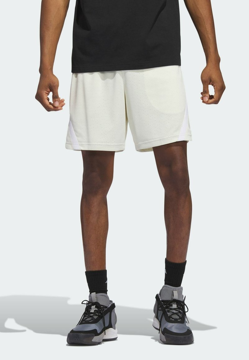 Homme portant un short de sport en mesh blanc, des chaussettes noires, des baskets grises et noires, et un t-shirt noir, debout avec les bras détendus le long du corps.