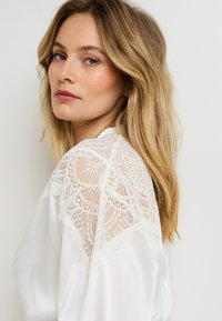 Blusa bianca con dettagli in pizzo intricati sulle spalle, texture liscia, vestibilità rilassata e una leggera lucentezza su tutto il tessuto.