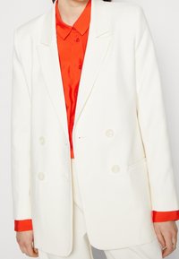 Vit dubbelknäppt blazer med slag. Bärs över en orange sidentopp med knapplås och en strukturerad passform.