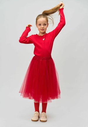 Haut à manches longues rouge côtelé avec un petit détail en forme de cœur, associé à une jupe en tulle rouge superposée, des chaussettes à pois rouges et des chaussures légères.