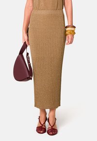 Goldene gerippte Strickrock mit slim, knöchellangem Design. Kombiniert mit roten Riemchenschuhen und einer burgunderfarbenen Handtasche. Glatte Textur, eleganter Abschluss.