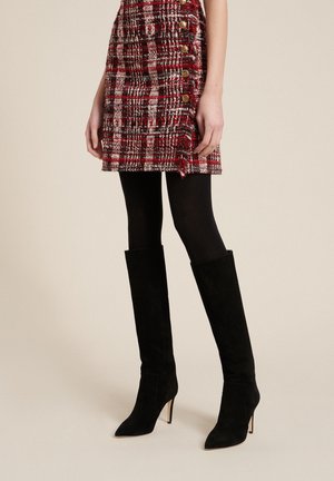 Kobieta ubrana w czerwoną kratowaną spódnicę na guziki, czarne rajstopy oraz czarne zamszowe kozaki na obcasie sięgające kolan, stojąca na beżowym tle.