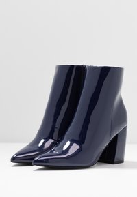 Botines de charol azul marino con punta afilada y tacón cuadrado, que cuentan con una cremallera lateral y un acabado brillante.