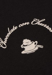 Texte blanc "Chocolate con Churros" se courbe au-dessus d'une tasse blanche fumante avec de la crème fouettée, posée sur une petite assiette, sur un fond en tissu noir.