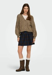 Strikket beige cardigan med V-hals og struktureret mønster, parret med en mørk ternet skaternederdel. Brune knæhøje støvler og hvide sokker.