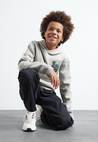 Sudadera gris con un bolsillo frontal y un logo verde, pantalones negros de corte holgado, y zapatillas blancas con acentos coloridos. El sujeto está de rodillas.