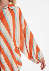 Blouse rayée orange et crème avec des accents bleus, en tissu léger, coupe ample, et manches retroussables, avec un col à boutons.