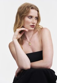 Gold pendant necklace with a circular medallion featuring an embossed letter. Model wearing a black strapless top, light skin, and wavy hair.