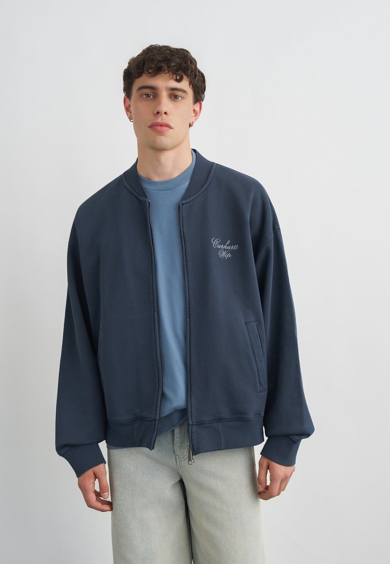 Νέος άνδρας που φοράει χαλαρό navy bomber jacket με λευκό λογότυπο σε σενάριο στο στήθος, ανοιχτό μπλε πουκάμισο από κάτω και ανοιχτόχρωμα τζιν, όρθιος.