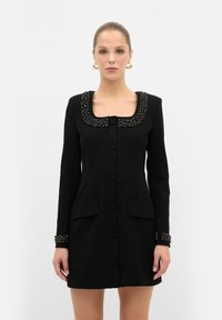 Robe noire avec un décolleté carré, col et poignets embellis, manches longues et boutons sur le devant. Comprend deux poches avant.