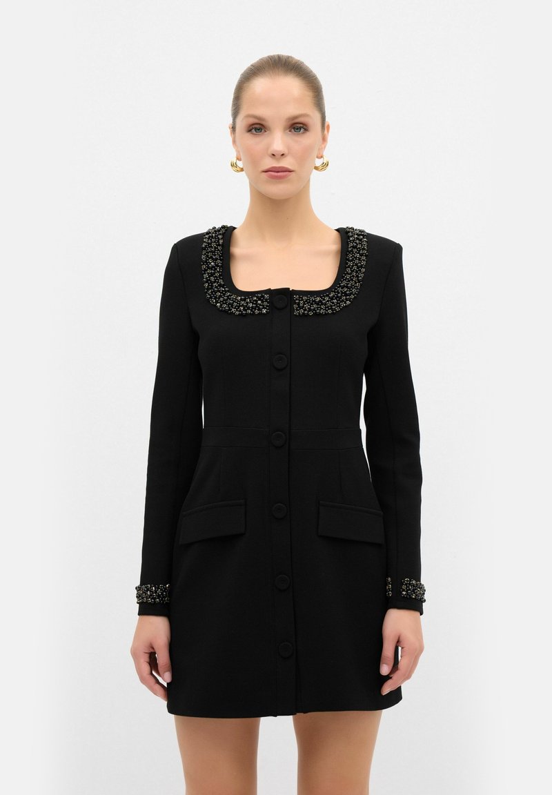 Robe noire avec un décolleté carré, col et poignets embellis, manches longues et boutons sur le devant. Comprend deux poches avant.