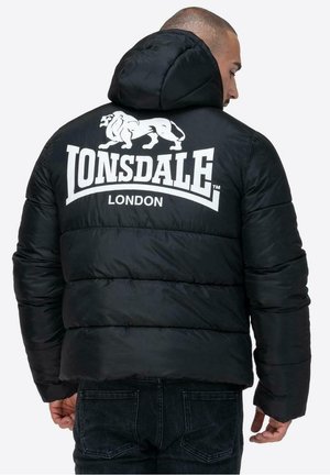 Man wearing black puffer jacket with hood, featuring large white "Lonsdale London" logo and lion graphic on the back, facing sideways.