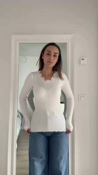 Femme portant un haut blanc côtelé à manches longues avec un col en dentelle et un jean bleu à jambes larges, debout dans un encadrement de porte à l'intérieur.