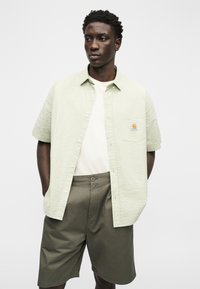 Άνδρας με σκούρο δέρμα και σγουρά μαλλιά, φοράει ανοιχτοπράσινο πουκάμισο με κοντά μανίκια πάνω από λευκή κοντομάνικη μπλούζα και χακί σορτς, στέκεται με τα χέρια στις τσέπες.