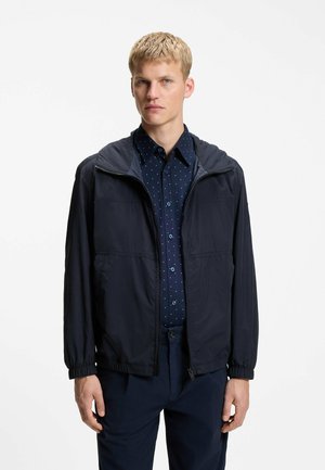 Jeune homme portant une veste zippée bleu marine sur une chemise bleu foncé à pois avec boutons et un pantalon bleu marine assorti, debout devant un fond blanc.