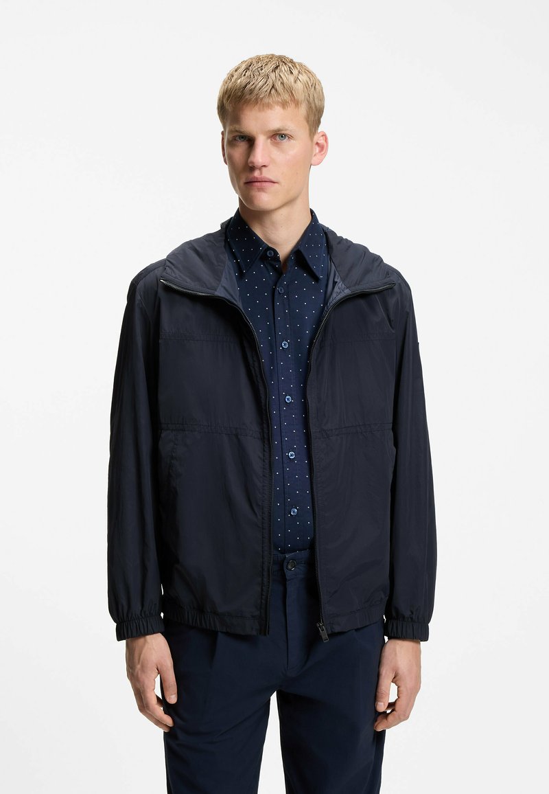 Jeune homme portant une veste zippée bleu marine sur une chemise bleu foncé à pois avec boutons et un pantalon bleu marine assorti, debout devant un fond blanc.