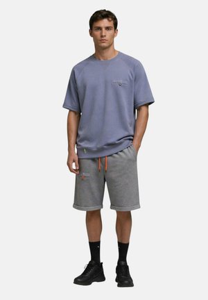 Jeune homme debout portant une chemise bleue à manches courtes, un short gris avec des cordons orange, des chaussettes noires et des baskets noires.