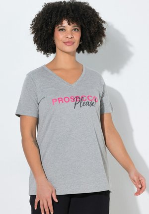 Kobieta o kręconych włosach, ubrana w szarą koszulkę z dekoltem w serek z napisem „PROSECCO Please!” w różowym i czarnym kolorze, stojąca na białym tle.