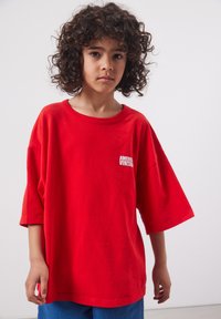 Enfant aux cheveux bouclés portant un T-shirt rouge surdimensionné avec le texte "American Vintage", debout devant un fond gris clair uni.