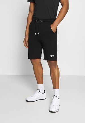 Mężczyzna ubrany w czarne szorty ze sznurkiem z logo Alpha Industries, białe sneakersy Nike i białe skarpety, stojący na jednolitym tle.