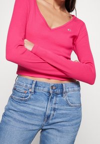 Růžová úzká dlouhá halenka s výstřihem do V a malým logem, spárovaná se světle modrými džínami s pěti kapsami a měděnými akcenty.