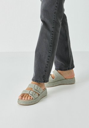 Grå flettede platåsandaler med dobbelt spænde og åbne sider, kombineret med mørkegrå jeans. Glat sål og tekstureret overdel.