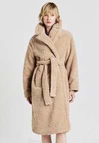 Accappatoio di peluche beige con colletto sciallato, cintura, due tasche laterali e tessuto testurizzato. Lunghezza al ginocchio con maniche lunghe.