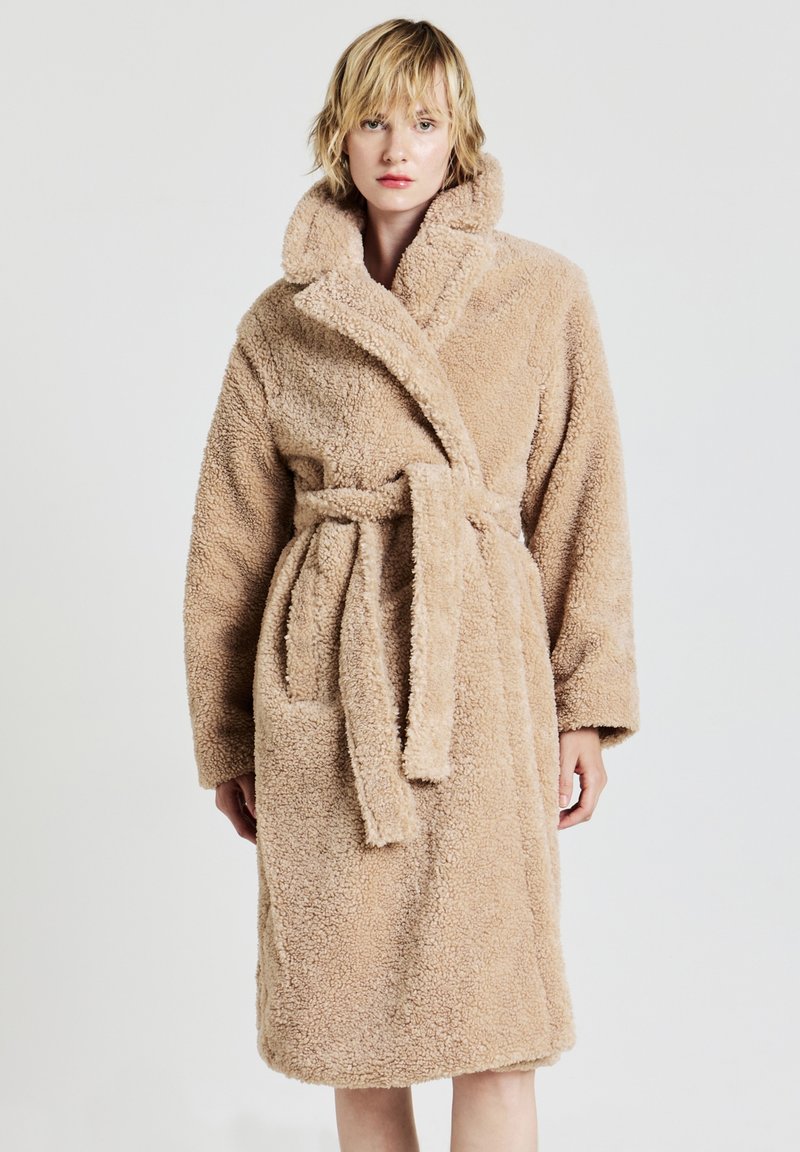 Accappatoio di peluche beige con colletto sciallato, cintura, due tasche laterali e tessuto testurizzato. Lunghezza al ginocchio con maniche lunghe.