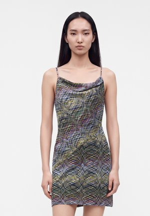 Jeune femme aux longs cheveux noirs et raides, portant une robe sans manches multicolore à motifs avec de fines bretelles, sur un fond clair uni.