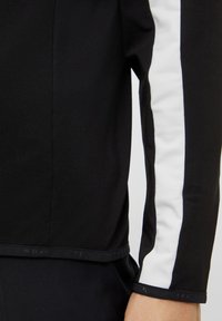 Chemise noire à manches longues avec une bande blanche sur l'épaule, tissu doux, et un design ajusté avec un ourlet droit.